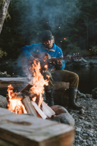 Man in blue shirt with black hat playing acoustic guitar Infront of campfire.