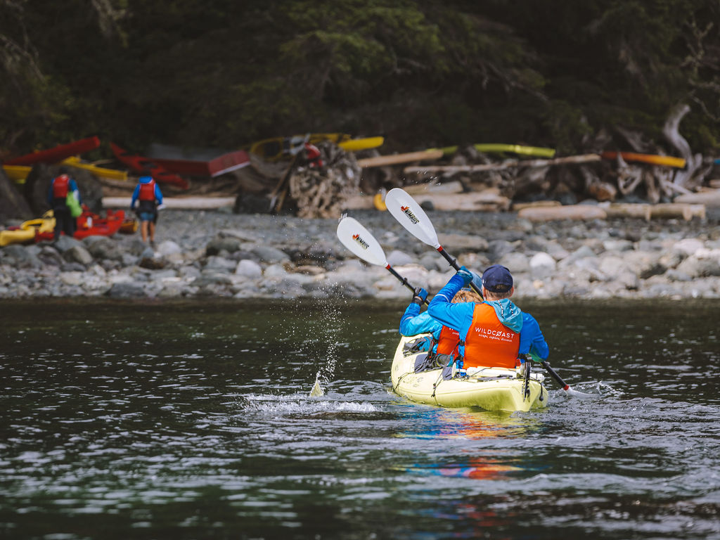 2 people in blue coats with red lifejackets paddling a yellow double kayak towards a rocky beach.