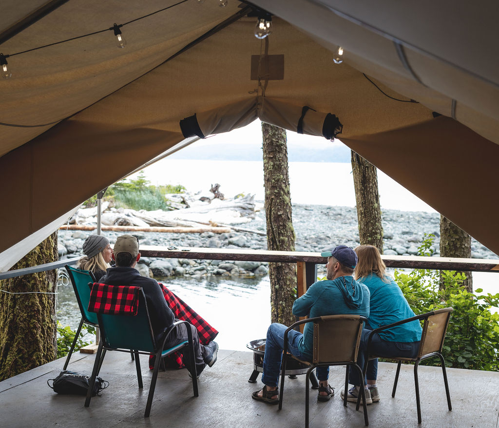 White tent with 2 men and 2 women sitting in camp chairs looking out at the ocean.