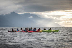Several multi colored kayaks rafted together in small ocean waves. Mountains in the background with sunlight shining through clouds.