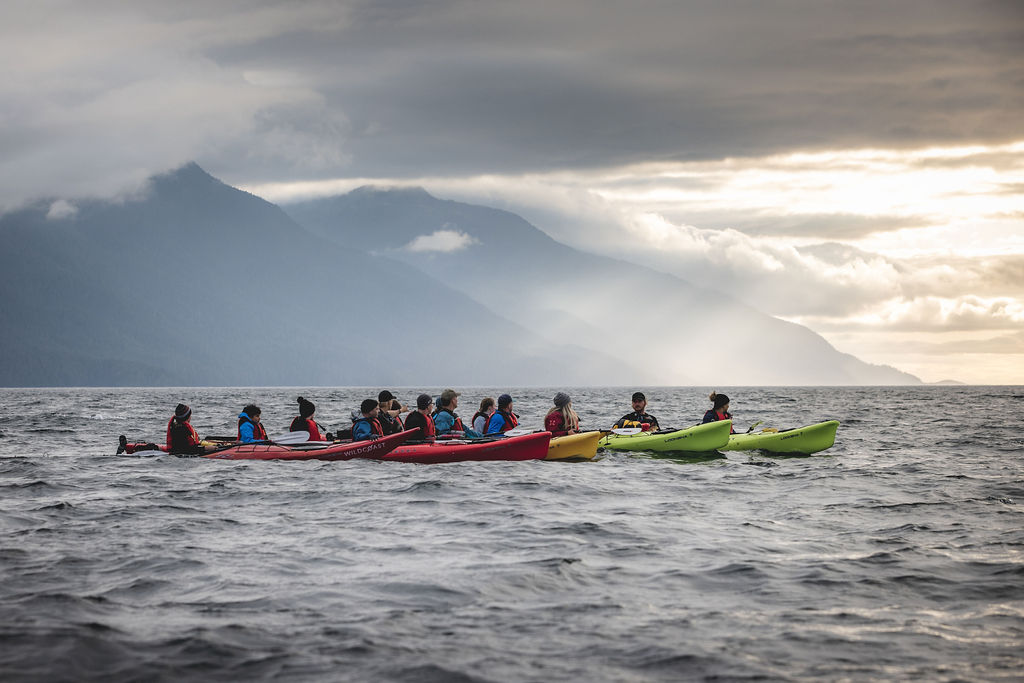 Several multi colored kayaks rafted together in small ocean waves. Mountains in the background with sunlight shining through clouds.