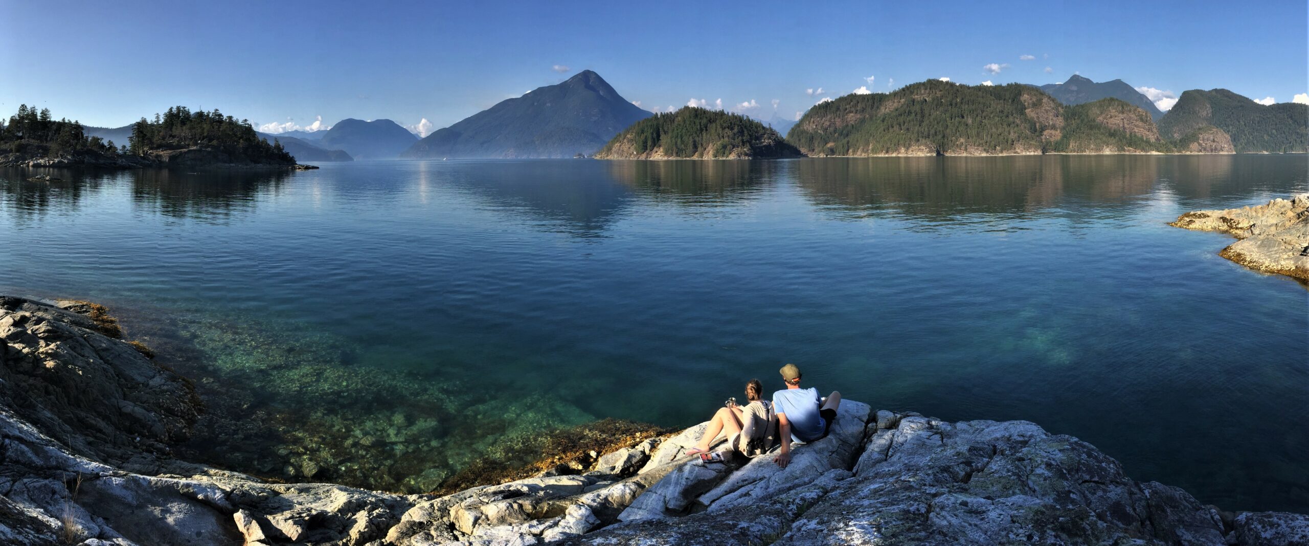 two people sitting on the edge of a cliff looking out at an island