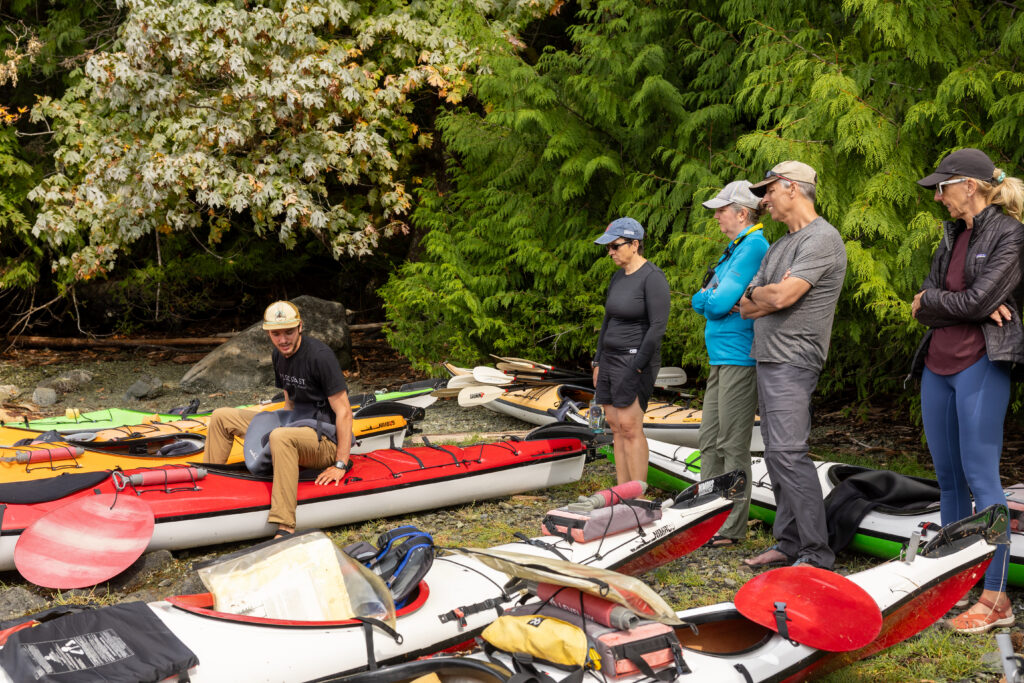 Guests getting a kayak briefing from their guide on a beach lined with sea kayaks