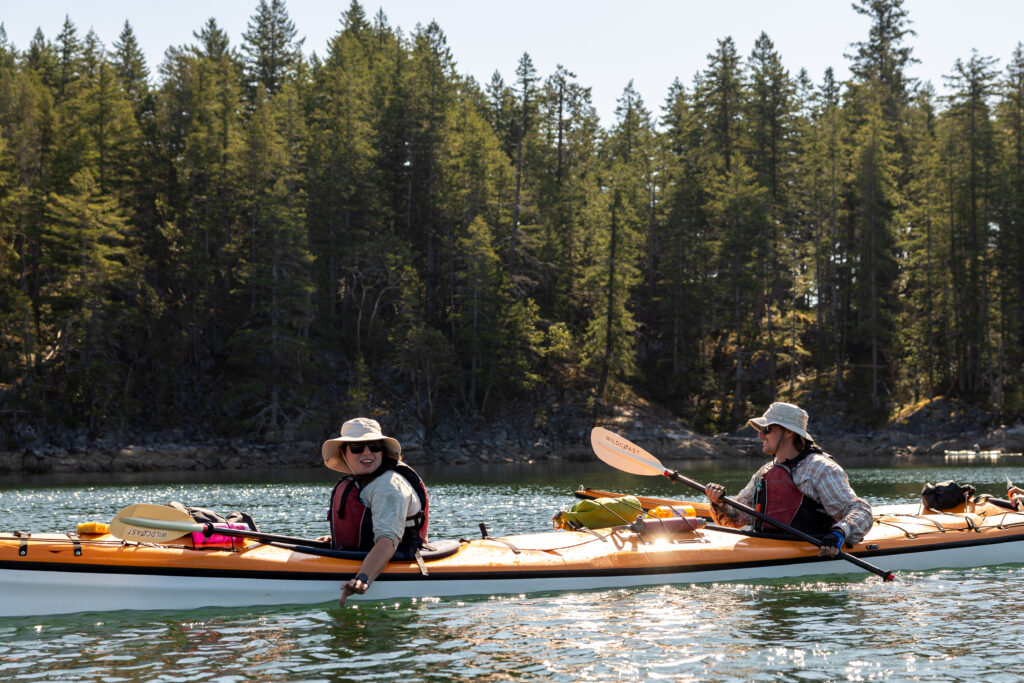 Two sea kayakers in a tandem kayak paddling past a forested shoreline