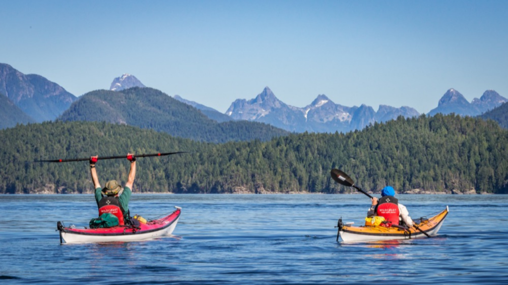 two people in kayaks paddling on the water with mountains in the background
